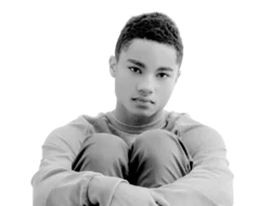 Black and white image of young man sitting with his knees up to his chest and his arms wrapped around his legs.