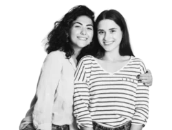 Two women pose for a picture together, smiling at the camera. The one on the left has curly dark hair and has her arm around the woman on the right who has straight dark hair and is wearing a striped shirt.