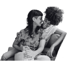 Two people sitting close and embracing each other on a couch. The person on the left has fair skin and long straight hair with a side undercut and the person on the right has darker skin with tight curls.