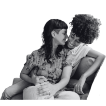 Two people sitting close and embracing each other on a couch. The person on the left has fair skin and long straight hair with a side undercut and the person on the right has darker skin with tight curls.